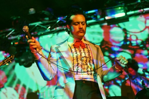 Lead singer of the band Creatures holding up a microphone, psychedelic lit up visuals behind him