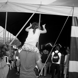 A man with a young girl on his shoulders watching a band from the back of a festival tent