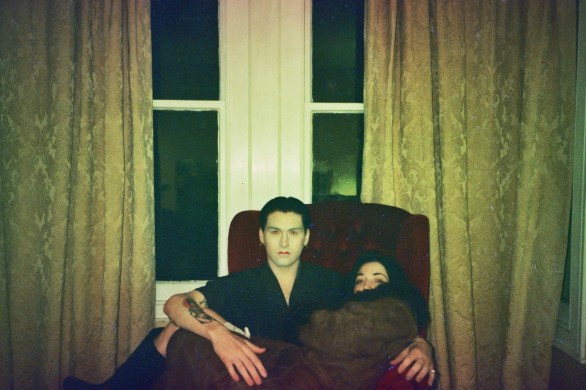 Will and Ellie who make up the band Robbie and Mona, sitting on a red armchair in front of a large window with heavy curtains