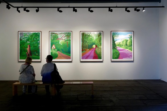 The silhouettes of the backs of two ladies sitting on a bench in an art gallery looking at 4 brightly coloured David Hockney pictures