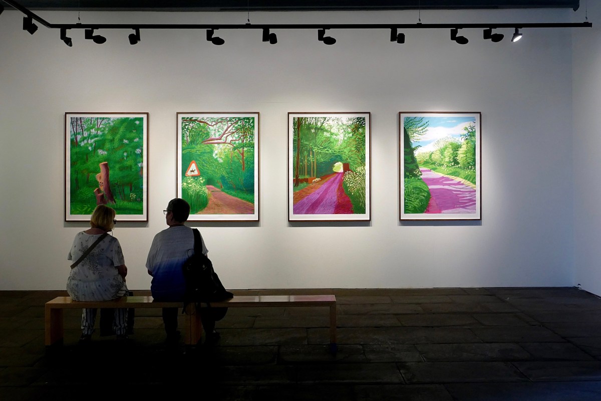 The silhouettes of the backs of two ladies sitting on a bench in an art gallery looking at 4 brightly coloured David Hockney pictures